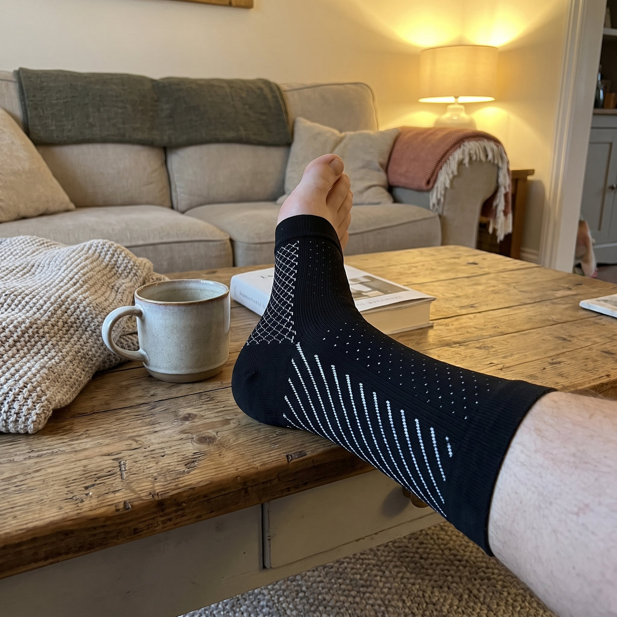 A black compression sock worn with the foot propped on a wooden table beside a mug