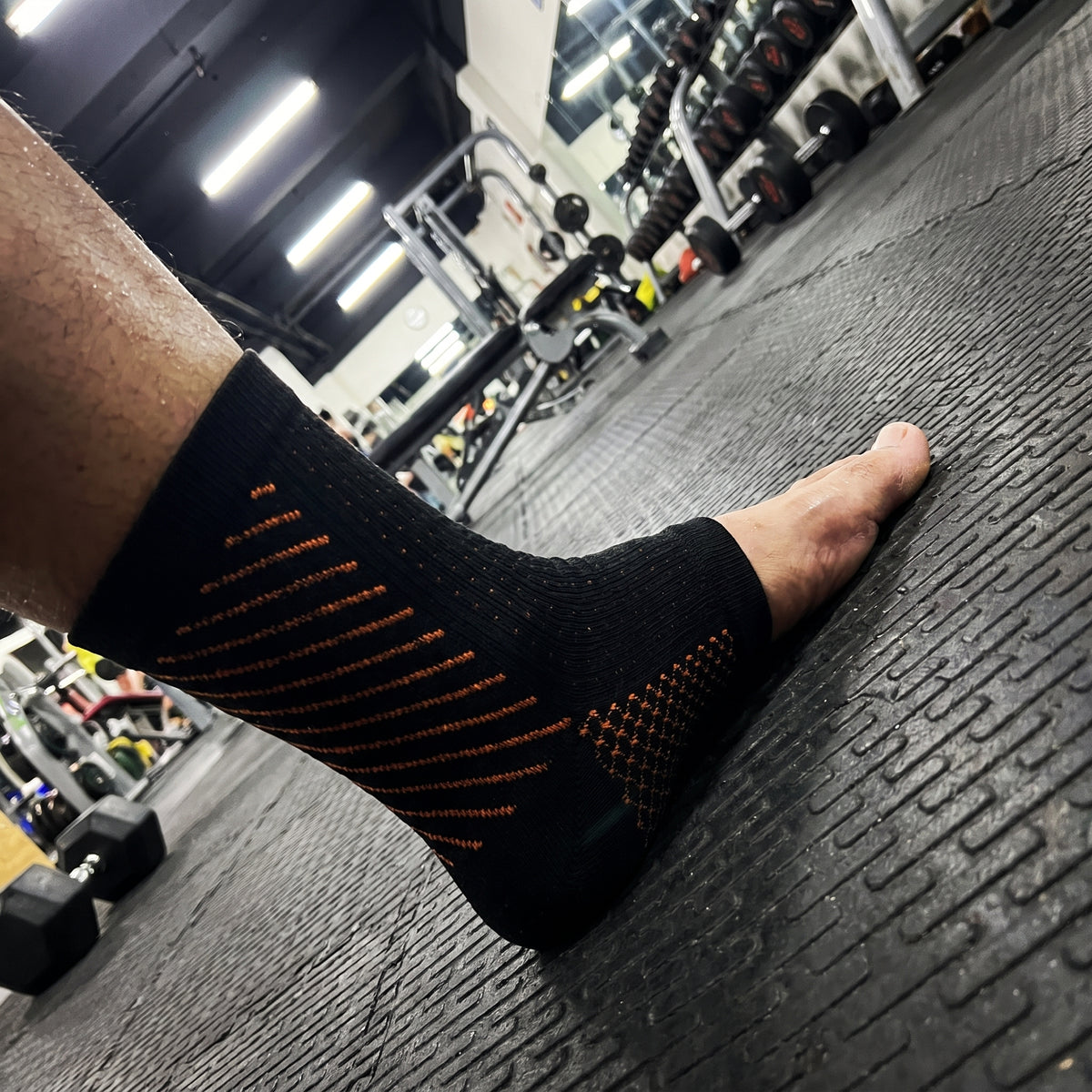 A black compression sock with orange arch stitching, worn on a gym floor