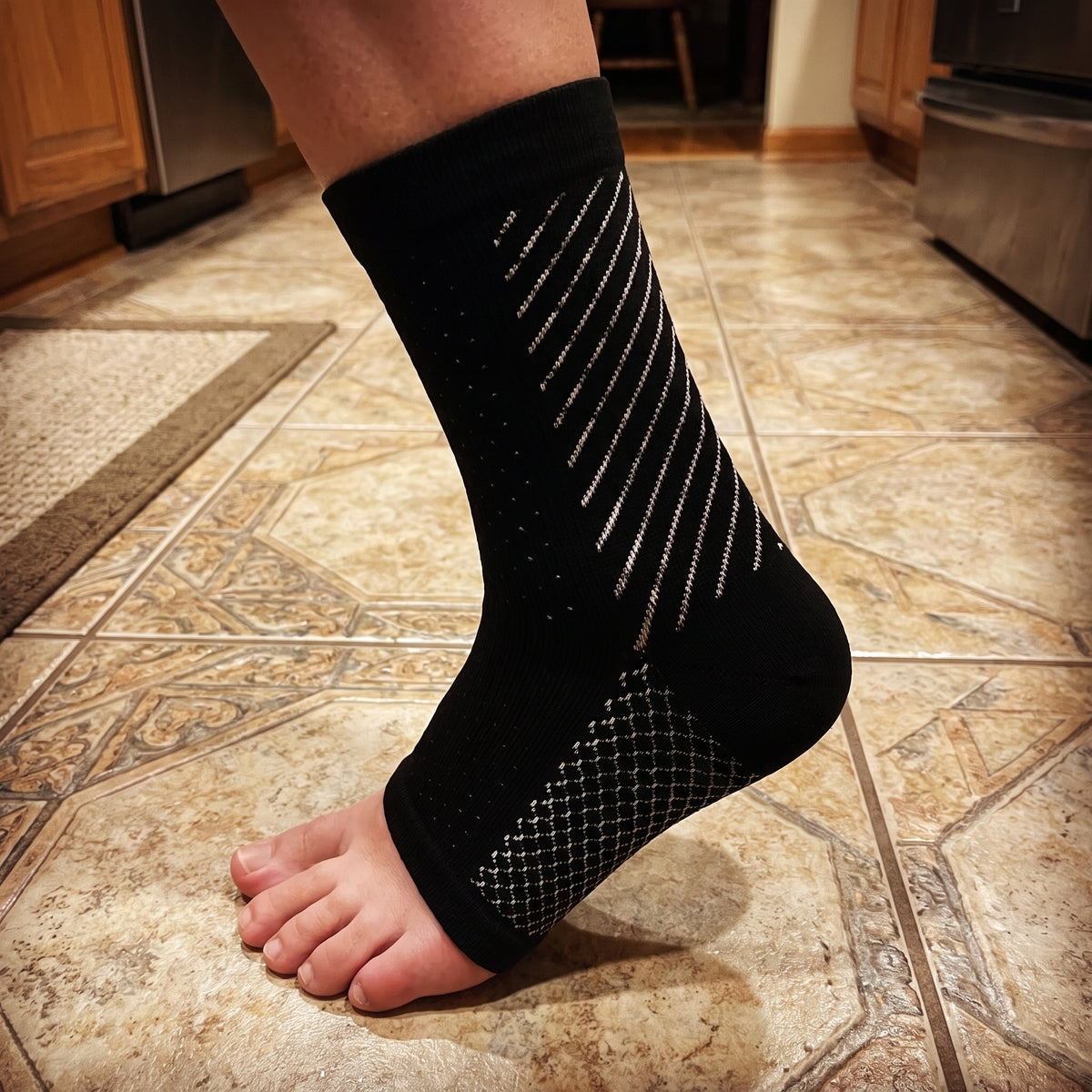 A black compression sock with white arch stitching, worn standing on a tiled kitchen floor