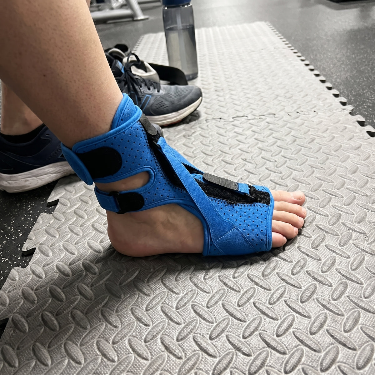The blue night splint fastened over a bare foot, worn while sitting on a gym mat