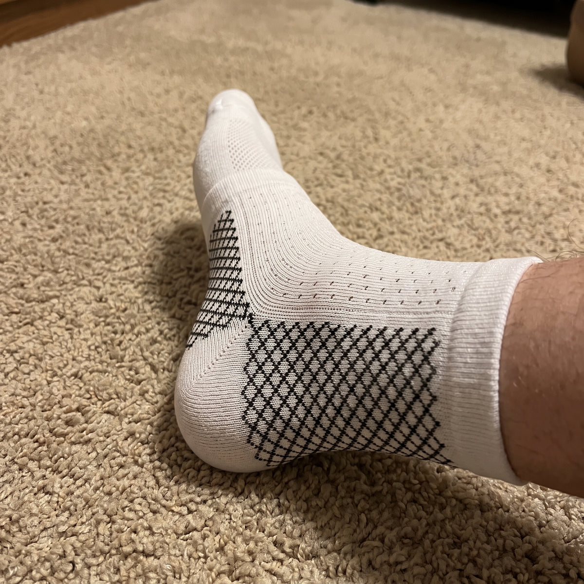 A white compression sock with black mesh panels, worn with the foot resting on carpet